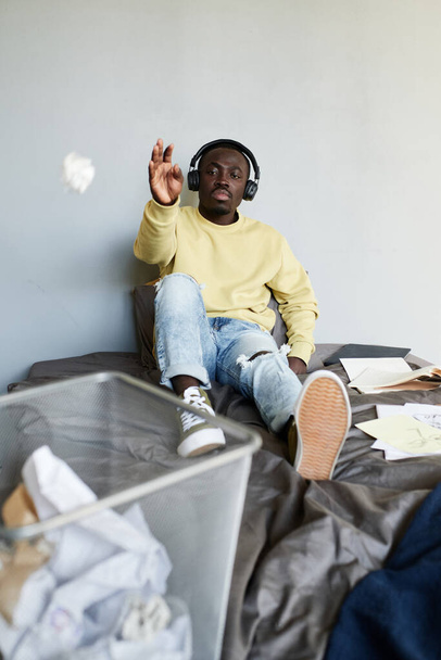 Lazy African-American student boy in wireless headphones sitting on bed and throwing crumpled paper into trash can while wasting time - Foto, Bild