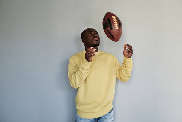 Serious young black rugby player of college team standing against gray wall and throwing ball - Valokuva, kuva