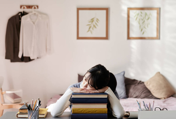 Exhausted Asian student girl sitting at table in room and sleeping on stack of books - Photo, image