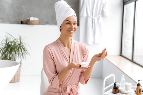 Hautreinigung. Schöne kaukasische brünette langhaarige Frau im Bademantel mit Handtuch auf dem Kopf, hält ein weißes Baumwollkissen, um ihre empfindliche Haut zu reinigen, verwendet Hautpflegeprodukte, während sie im Badezimmer sitzt - Foto, Bild
