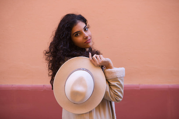 Young and beautiful Hispanic brunette woman with curly hair plays with a hat on a pink background. The woman plays hide and seek behind the hat making different funny expressions. - Photo, Image
