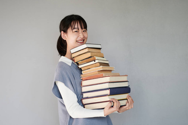 Retrato de estudante asiático positivo menina de pé com pilha de livros contra fundo cinza - Foto, Imagem
