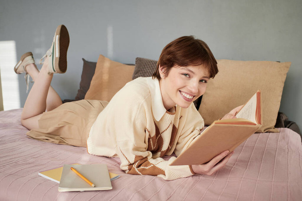Porträt eines glücklichen attraktiven Studentenmädchens mit roten Haaren, das auf dem Bett liegt und Lehrbücher liest - Foto, Bild