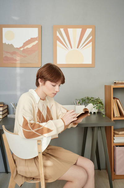 Serious redhead girl in beige outfit sitting at desk in students room and making notes in sketchpad - 写真・画像