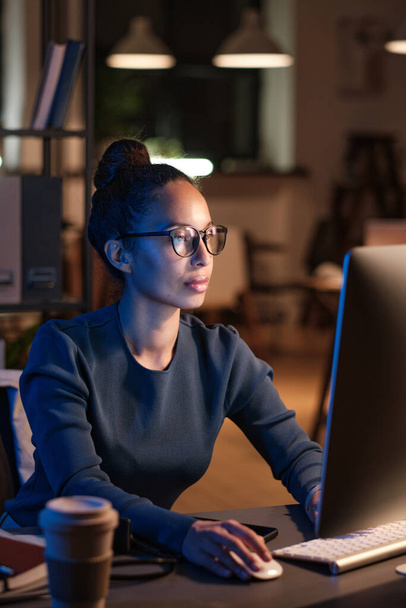 Concentrated beautiful multiethnic businesswoman in glasses sitting at desk in dark office and working with online documents on computer - Foto, immagini