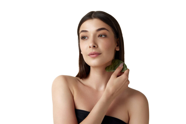 Beautiful young woman with well-kept healthy skin taking care after face with cosmetological massage tool against white background. Concept of natural female beauty, body and skincare, cosmetology, ad - Zdjęcie, obraz
