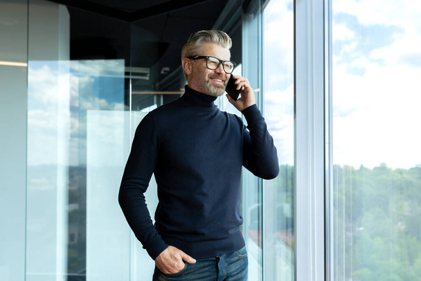 Senior male businessman, designer stands by the window in the office center and prays on the phone. A smiling man looks out the window, keeps his hand in his pocket. - Foto, afbeelding