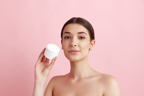 Portrait of young beautiful woman posing with face moisturizing cream against pink studio background. Organic cosmetics. Concept of natural female beauty, body and skincare, cosmetology, health, ad - Fotó, kép