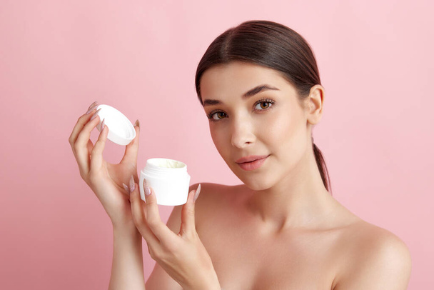 Portrait of young beautiful woman posing with face moisturizing cream against pink studio background. Skin hydration. Concept of natural female beauty, body and skincare, cosmetology, health, ad - Valokuva, kuva
