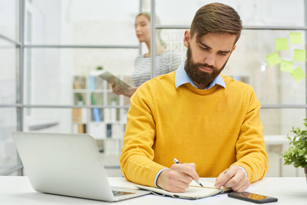 Serious busy handsome young businessman in yellow sweater writing ideas in diary while working in modern office - Φωτογραφία, εικόνα