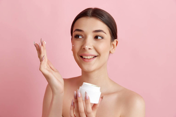 Cream with spf care. Portrait of young beautiful woman posing with face moisturizing cream against pink studio background. Concept of natural female beauty, body and skincare, cosmetology, health, ad - Фото, изображение