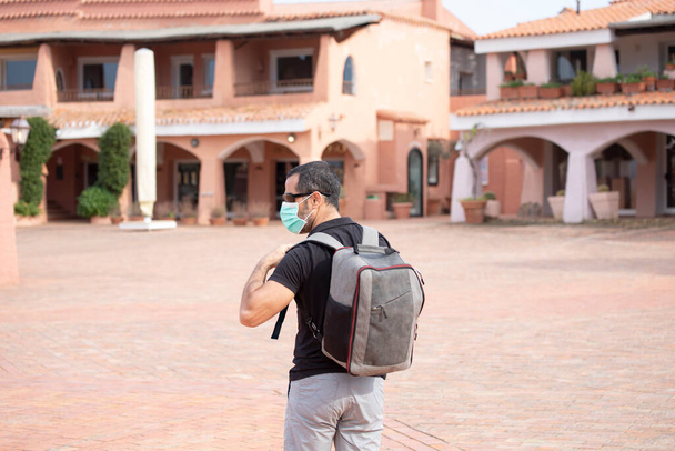 Hombre de pelo oscuro en traje de verano y mochila pasea por una ciudad turística con máscara facial protectora - Foto, imagen