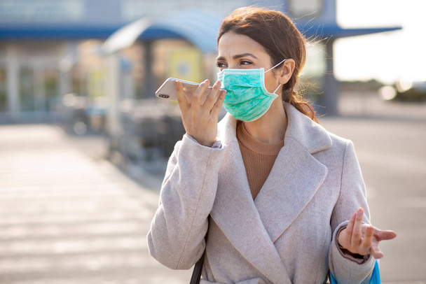 girl with coat and facial surgical mask, stops in front of the supermarket with the shopping bag and speaks on a cell phone - Photo, Image