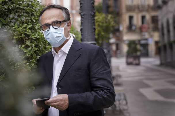 portrait of middle-aged man in jacket with protective face mask, standing on a sidewalk with public greenery behind - Foto, afbeelding