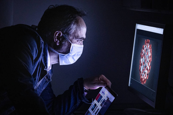hombre con una máscara y una computadora en su lugar de trabajo por la noche - Foto, imagen