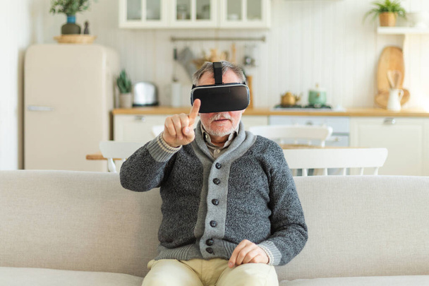 Opgewonden volwassen senior man dragen met behulp van virtual reality metaverse VR bril headset thuis. Grootvader die lucht aanraakt tijdens VR-ervaring op virtual reality helm. Simulatie high-tech videogame - Foto, afbeelding