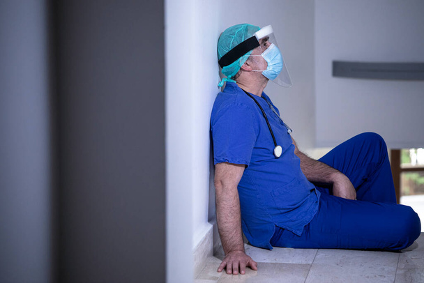 lääkäri sinisessä takissa, jolla on suoja ja kasvokilpi, tuhoutunut väsymyksestä sairaalaosastolla - Valokuva, kuva