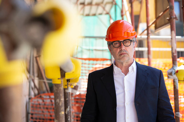 ingeniero con casco, naranja, chaqueta y gafas se encuentra en medio de un sitio de construcción con andamios - Foto, imagen