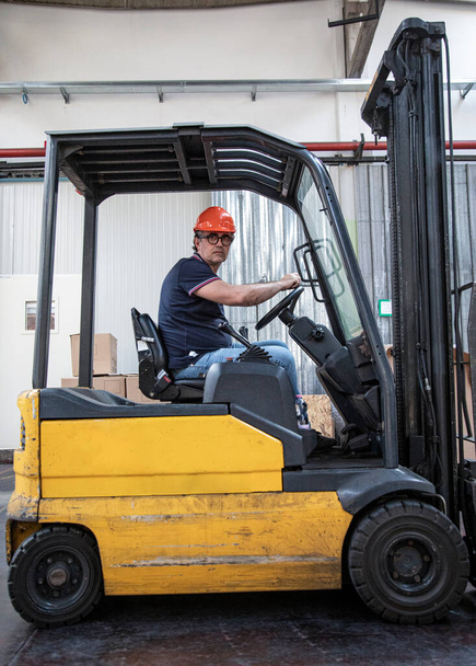 Worker with protective helmet and black glasses is driving a forklift truck inside a warehouse - Photo, Image