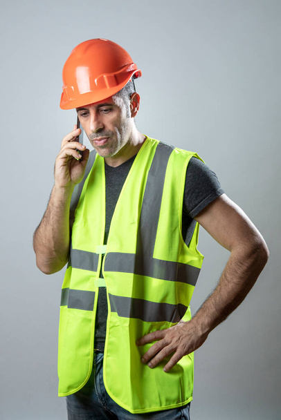 munkás narancssárga keménykalapban és kézi sárga mellényben telefonál mobiltelefonnal, szürke alapon elszigetelve - Fotó, kép