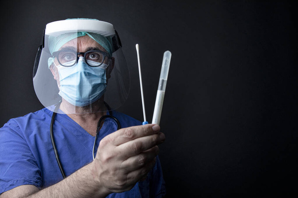 Врач с хирургической защитой и синим лабораторным халатом показывает тампон, выделенный на черном фоне - Фото, изображение