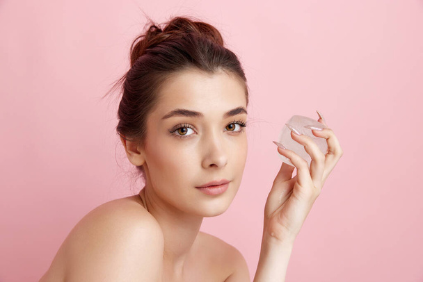 Young beautiful woman taking care after her perfect skin with ice cube over pink studio background. Refreshment and anti-wrinkle care. Concept of natural female beauty, skincare, cosmetology, health - Φωτογραφία, εικόνα