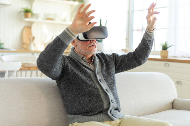 Excited mature senior man wearing using virtual reality metaverse VR glasses headset at home. Grandfather touching air during VR experience on virtual reality helmet. Simulation hi-tech videogame - Fotografie, Obrázek