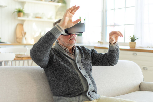 Ενθουσιασμένος ώριμος ηλικιωμένος που φοράει γυαλιά εικονικής πραγματικότητας στο σπίτι. Παππούς αγγίζοντας αέρα κατά τη διάρκεια VR εμπειρία σε κράνος εικονικής πραγματικότητας. Προσομοίωση βιντεοπαιχνιδιού υψηλής τεχνολογίας - Φωτογραφία, εικόνα