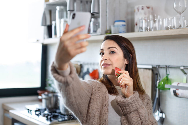Bruneta dívka vezme selfie s mobilním telefonem při jídle jahody v kuchyni - Fotografie, Obrázek