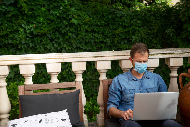 een man met een laptop en een masker thuis in de tuin - Foto, afbeelding