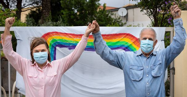 älteres Paar mit Operationsmaske, lässig gekleidet vor einer Regenbogenzeichnung an einem Tor vor ihrem Haus, zärtlich Händchen haltend. - Foto, Bild
