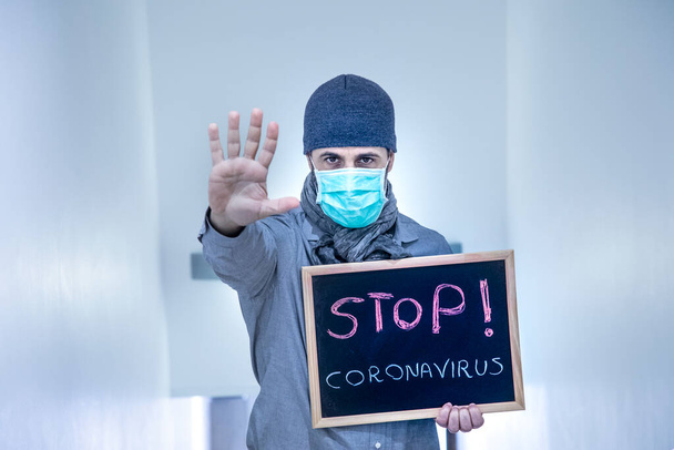 Homem com boné na cabeça e máscara, isolado em um fundo claro, segura um quadro negro com as palavras "stop Coronavirus" - Foto, Imagem