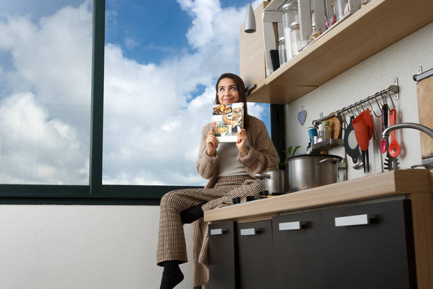 casual ragazza vestita cerca di trovare una buona ricetta seduta sulla parte superiore del suo piano di lavoro della cucina di fronte a una grande finestra con il cielo blu sullo sfondo - Foto, immagini