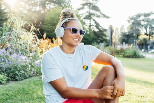 Smiling mixed race young woman with a rainbow badge, symbol of LGBTQ social community listening music with wireless headphones while relax in park. Social Diversity acceptance, feel free lifestyle - Foto, immagini