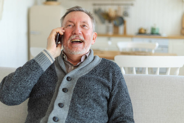 Mutlu orta yaşlı bir adam aile dostlarıyla akıllı telefondan konuşuyor. Yetişkin bir büyükbaba cep telefonuyla yetişkin çocuklarla konuşuyor, evde dinleniyor. Eski nesil modern teknoloji kullanımı - Fotoğraf, Görsel