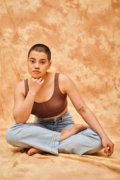 body confidence, acceptance, curvy young and tattooed woman in jeans and crop top sitting with crossed legs on mottled beige background, personal style, self-acceptance, generation z, denim fashion - Fotó, kép