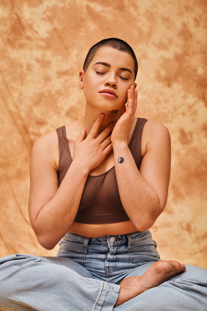 body image, relaxation, curvy young and tattooed woman in jeans and crop top sitting with crossed legs on mottled beige background, closed eyes, personal style, self-acceptance, generation z  - Photo, Image