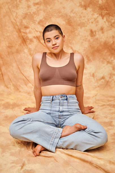 body positivity, casual attire, curvy young and tattooed woman in jeans and crop top sitting with crossed legs on mottled beige background, personal style, self-acceptance, generation z  - Foto, afbeelding
