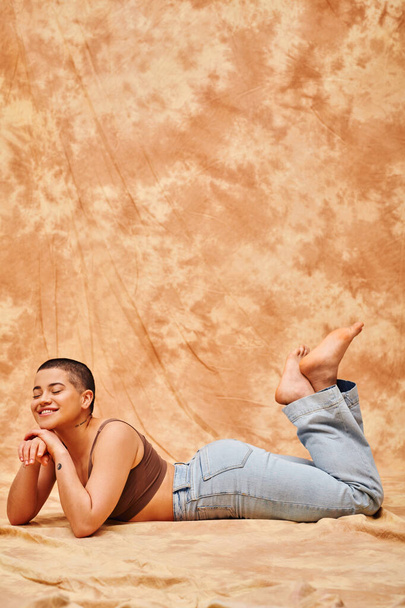 body positivity, representation of body, curvy and tattooed woman in jeans and crop top lying on mottled beige background, smiling with closed eyes, denim fashion, personal style, generation z  - Photo, Image