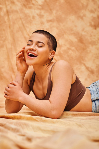 body positivity, curvy and excited woman in crop top posing with hand near face on mottled beige background, tattooed, representation of body, different body shapes, generation z, youth  - Φωτογραφία, εικόνα