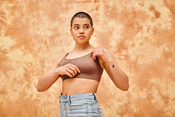 body positivity movement, curvy young woman with tattoos posing in jeans and crop top on mottled beige background, representation of body, confidence, casual attire, generation z, looking away - Photo, Image