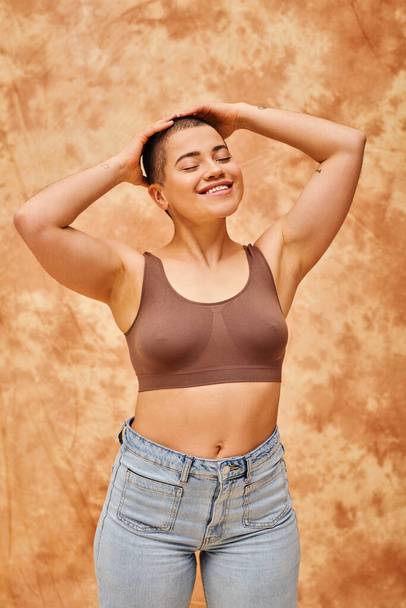 body acceptance, curvy and cheerful woman in crop top posing on mottled beige background, smiling with closed eyes, representation of body, different shapes, generation z, youth, tattooed   - Foto, Bild