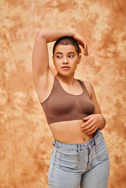 body love, curvy young woman in crop top posing with raised hand on mottled beige background, looking away, representation of body, different shapes, generation z, youth, tattooed, relaxed pose  - Zdjęcie, obraz