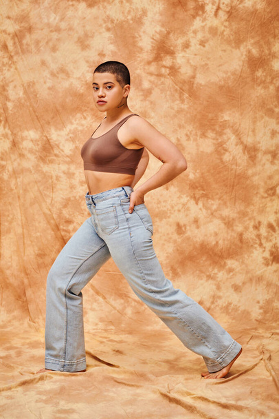 Jeansmode, Generation Z, junge kurvige Frau mit Tätowierungen posiert mit den Händen in den Taschen auf fleckigem beigen Hintergrund, verschiedene Formen, Body Positivity Bewegung, Selbstwertgefühl, Selbstvertrauen - Foto, Bild