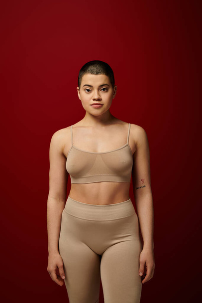 body diversity, young tattooed woman in beige underwear posing on red background, body positivity, real people, burgundy, comfortable in skin, curvy model, generation z, self love, looking at camera  - Zdjęcie, obraz
