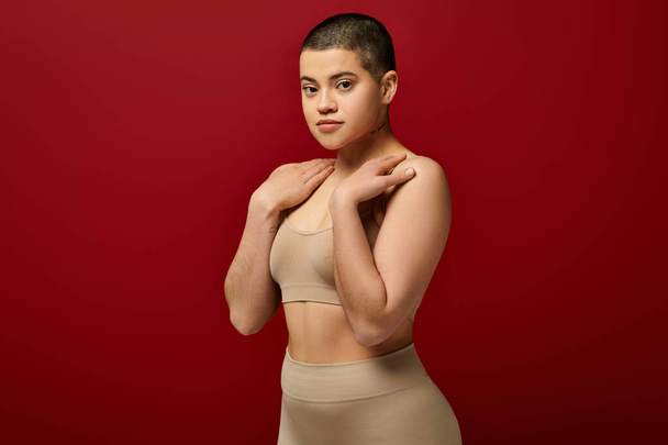 body appearance, young tattooed woman in beige underwear posing on red background, body positivity, natural curves, comfortable in skin, curvy model, generation z, self love, looking at camera  - Foto, afbeelding