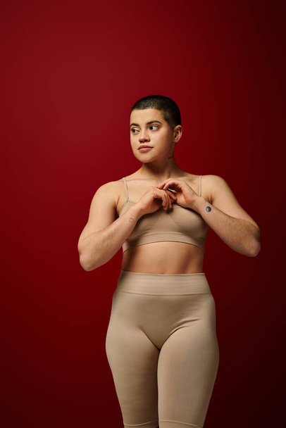 Body Empowerment, junge tätowierte Frau in beiger Unterwäsche posiert auf rotem Hintergrund, Körperpositivität, kurvige Mode, bequem in der Haut, kurviges Modell, Generation z, Selbstliebe, Wegschauen - Foto, Bild
