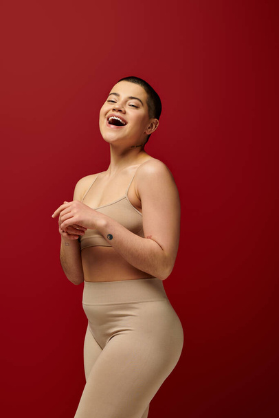 self acceptance, amazed and tattooed woman in beige underwear posing on red background, body positivity, curvy fashion, comfortable in skin, curvy model, generation z, self love, short hair  - Foto, afbeelding