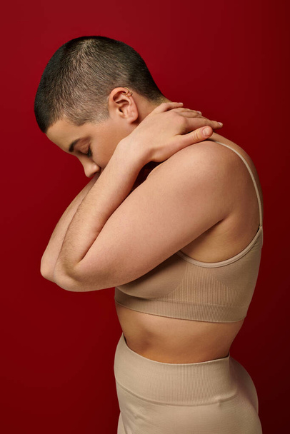 self acceptance, side view of young woman in beige underwear posing on red background, curvy fashion, comfortable in skin, self-acceptance, generation z, body diversity, short hair  - Фото, зображення