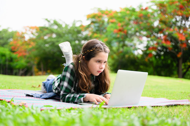 Glückliches Mädchen mit Laptop und Internet spielen im Gartenpark am Wochenende, Kind entspannen mit Aktivitäten, Mädchen hören Musik und lesen Buch im Park, Kind mit Technologie lernen und Bildung - Foto, Bild